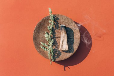 Burning palo santo stick with smoke on wooden stand over red orange background, burning incense. Holy wood sticks for meditation. Room fumigation ritual. Selective focus