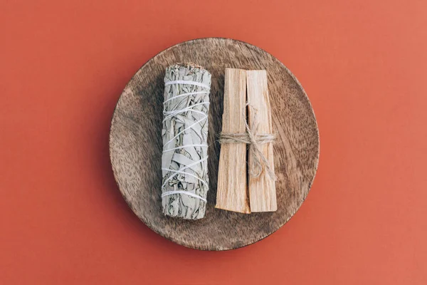 Incense bundle for meditation and room fumigation. White sage and palo santo sticks on wooden stand over orange red background. Holy wood sticks. Selective focus