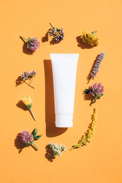 Herbal beauty products - tube with hand cream, face cream or body lotion with natural ingredients with flowers around over orange background. Flat lay style, mockup. Vertical image