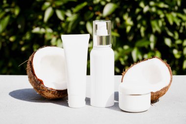 Coconut beauty products bundle. White bottles with coconut cosmetics - oil, body milk or lotion, cream jar against natural green leaves background. Natural cosmetic treatment for skincare. Mockup