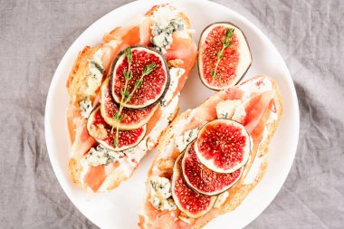 Taze kruvasan bruschetta, olgun incir, mavi peynir, prosciutto, ceviz ve beyaz tabakta bitkiler. Seçici odak