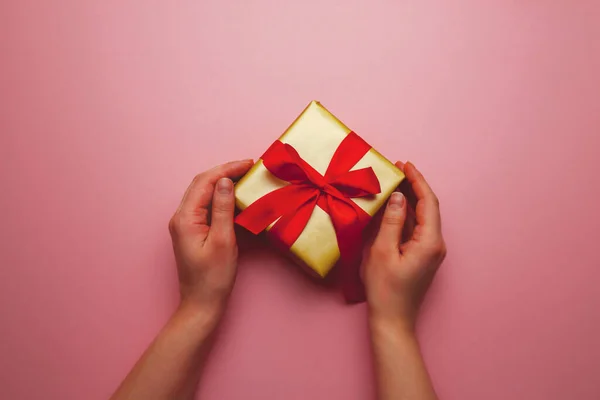 Dárek na dovolenou. Krabička ve zlatém dárkovém balení s červenou mašlí, na růžovém pozadí. Veselé Vánoce, Valentýne. Pohlednice s láskou. — Stock fotografie
