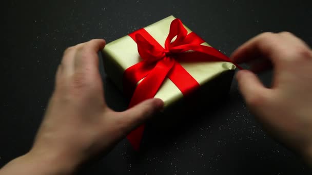 Het openen van een feestelijk geschenk verpakt in goud en een rode strik op een zwarte achtergrond. Een aangenaam cadeau en verrassing voor geliefden. — Stockvideo