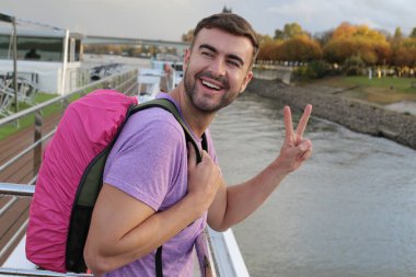 Sırt çantasıyla seyahat ederken barış jesti yapan yakışıklı bir gencin portresi