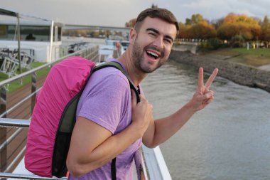 Sırt çantasıyla seyahat ederken barış jesti yapan yakışıklı bir gencin portresi