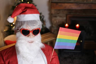 Noel Baba kostümü ve kalp şeklinde güneş gözlüğü ve Igbtq gökkuşağı bayrağı giymiş birinin portresi.