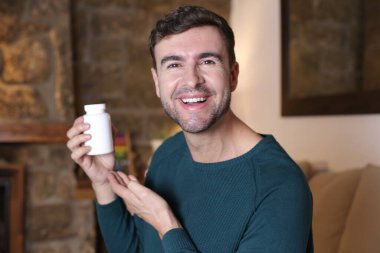 Evde elinde beyaz plastik ilaç ya da vitamin kavanozu tutan yakışıklı genç adamın yakın plan portresi.