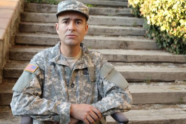 Parktaki merdivenlerin önünde tekerlekli sandalyede oturan üniformalı yakışıklı genç askerin yakın plan portresi.