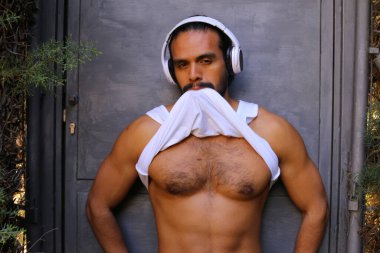 Kulaklıklı yakışıklı genç adamın yakın plan portresi gri duvar arkasında beyaz gömleğini çıkarıyor.