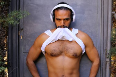 Kulaklıklı yakışıklı genç adamın yakın plan portresi gri duvar arkasında beyaz gömleğini çıkarıyor.