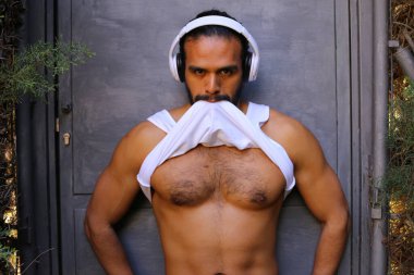 Kulaklıklı yakışıklı genç adamın yakın plan portresi gri duvar arkasında beyaz gömleğini çıkarıyor.