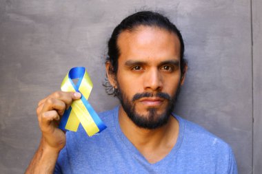 Gri duvar arkasında Ukrayna bayrağı renginde kurdele tutan yakışıklı genç adamın yakın plan portresi.