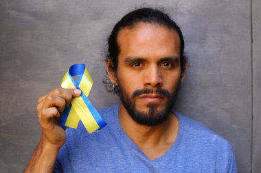 Gri duvar arkasında Ukrayna bayrağı renginde kurdele tutan yakışıklı genç adamın yakın plan portresi.