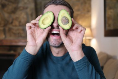 Evinde iki parça avokado tutan yakışıklı genç adamın yakın plan portresi.