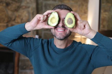 Evinde iki parça avokado tutan yakışıklı genç adamın yakın plan portresi.