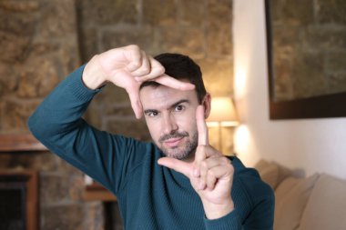 Yakışıklı genç adamın yakın plan portresi. Çerçeve jestini evde elleriyle gösteriyor.