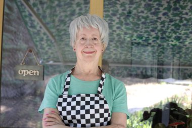 Kafe kapısında kareli önlüklü olgun bir kadının yakın plan portresi.