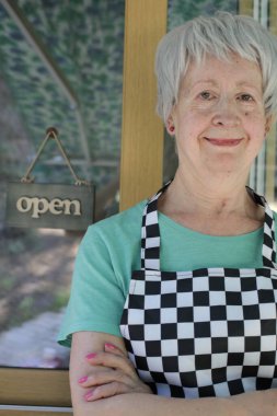 Kafe kapısında kareli önlüklü olgun bir kadının yakın plan portresi.