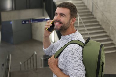 Sırt çantalı yakışıklı genç adamın yakın plan portresi tren istasyonunda telefonla konuşuyor.
