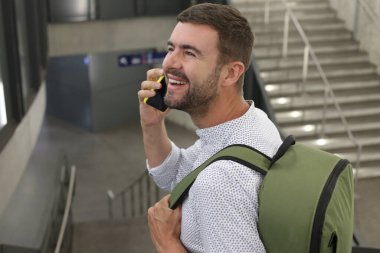 Sırt çantalı yakışıklı genç adamın yakın plan portresi tren istasyonunda telefonla konuşuyor.