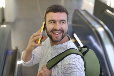 Sırt çantalı yakışıklı genç adamın yakın plan portresi tren istasyonunda telefonla konuşuyor.