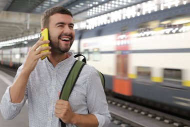 Sırt çantalı yakışıklı genç adamın yakın plan portresi tren istasyonunda telefonla konuşuyor.
