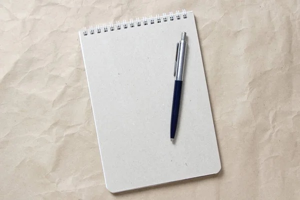 Gray notepad with white coiled spring and pen on a background of beige crumpled craft paper — Stock Photo, Image