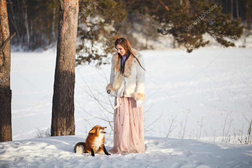 A beautiful girl walks with a fox in a snowy forest. Cold weather. Winter's Tale. Red fox. Photoshoot in a fairy-tale style.