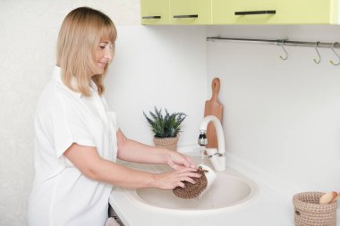 Bulaşıkları doğal sıfır atık süngeriyle yıkayan kadın og jute yaptı. El yapımı eko mutfak aşıları. Sarışın orta yaşlı kadın modern bir mutfakta temizlik yapıyor..