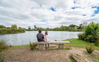 Auckland Yeni Zelanda 'da Auckland Gölü' nün önündeki Park Bench 'te oturan abla ve kardeş.