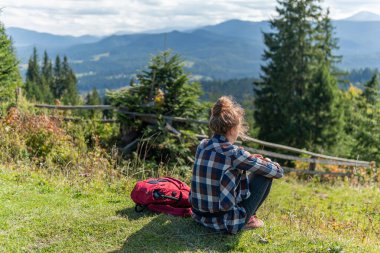 Genç bir kadın çimenlerde oturuyor, güneşli bir yaz gününde dağların tepesinde dinleniyor.