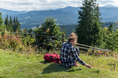 Genç bir kadın çimenlerde oturuyor, güneşli bir yaz gününde dağların tepesinde dinleniyor.