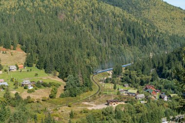 Parlak güneşli bir günde yeşil ormandan geçen bir tren ve küçük bir köyde evleri olan dağlardaki tren istasyonunda hava manzarası