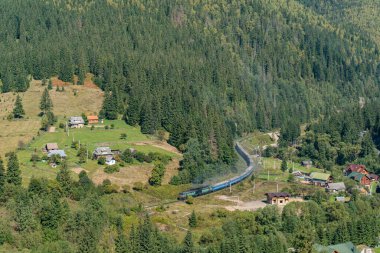 Parlak güneşli bir günde yeşil ormandan geçen bir tren ve küçük bir köyde evleri olan dağlardaki tren istasyonunda hava manzarası