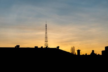 Gün batımında şehirdeki bir binanın çatısında çalışan iki adamın silueti. Altın saat, büyük kule ve konut binaları.