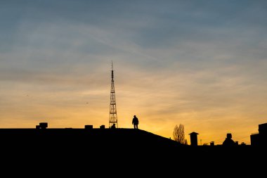 Gün batımında şehirdeki bir binanın çatısında çalışan iki adamın silueti. Altın saat, büyük kule ve konut binaları.