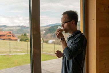 A man is standing at a window, staring out and drinking coffee. Window overlooks a beautiful view, landscape with mountains and fields