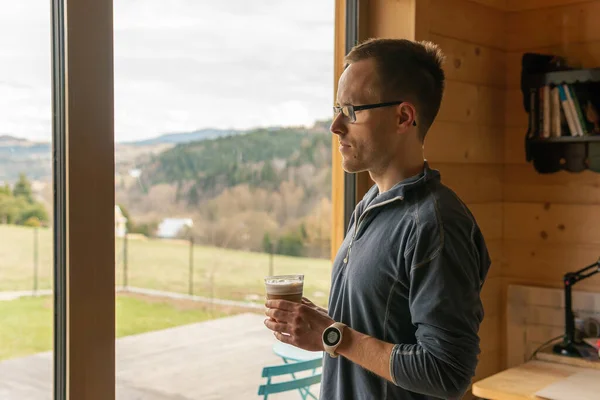 A man is standing at a window, staring out and drinking coffee. Window overlooks a beautiful view, landscape with mountains and fields