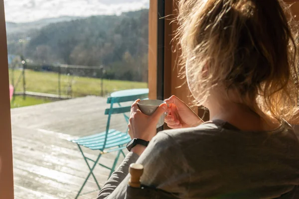 A woman is having morning tea or coffee, relaxing and looking at amazing view at mountains on a bright sunny day. Breathtaking view from a summer house.