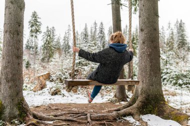 Poland, Rabka-Zdroj   April 10, 2022: A pregnant woman is swinging outdoors in winter with amazing view on the forest