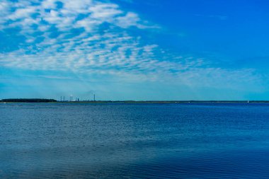 Su yüzeyinde dalgalanmalar, mavi gökyüzünde bulutlar. Tankın diğer tarafında dumanlı endüstriyel borular var..