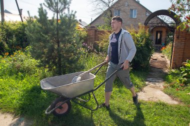 30 yaşında beyaz bir adam, yazlık evinde yolları betonla doldurmak için kum yüklemek için bir bahçe el arabası taşıyor.