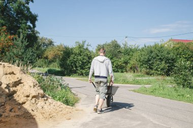 30 yaşında beyaz bir adam, yazlık evinde yolları betonla doldurmak için kum yüklemek için bir bahçe el arabası taşıyor.