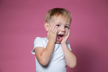 Beyaz çocuk, 3 yaşında. Pembe arka plandaki kameraya bakarken korkunç suratlar yapıyor.