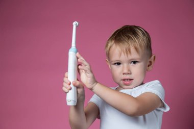Gülümseyen beyaz çocuk pembe arka planda elektrikli sonik diş fırçası tutuyor..