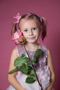 Pembe arka planda gül çiçeğiyle poz veren şenlikli elbiseli beyaz bir kız kameraya bakıyor.