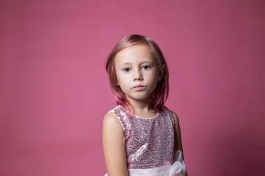 Pembe arka planda pulları olan şenlikli elbiseli beyaz bir kız kameraya bakıyor.