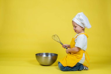 Küçük beyaz çocuk aşçıyı oynuyor, önlüklü çocuk ve yerde oturan aşçı şapkası sarı arka planda çırpıyor.