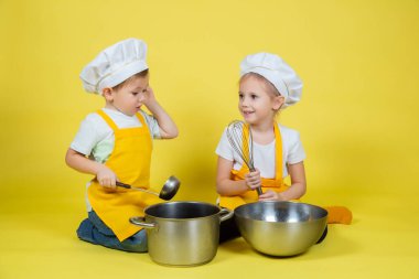 Küçük beyaz çocuklar şefçilik oynuyor, önlüklü çocuklar ve yerde oturan aşçı şapkası ve sarı arka planda çırpma sesi.