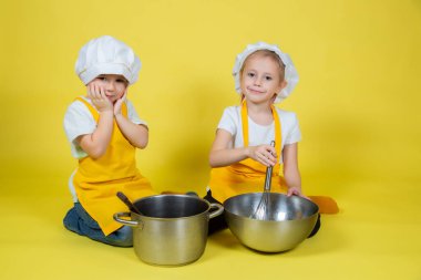 Küçük beyaz çocuklar şefçilik oynuyor, önlüklü çocuklar ve yerde oturan aşçı şapkası ve sarı arka planda çırpma sesi.